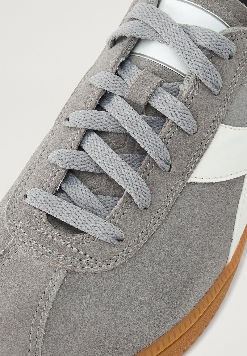 Sneaker en suede gris avec lacets gris, accents blancs et semelle en caoutchouc marron. Présente une texture lisse et de subtils détails de couture.