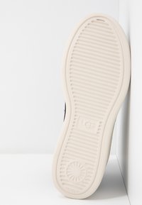 Černá podrážka boty s bílou gumovou podešví s žebrovanou strukturou a kruhovým designovým prvkem, uprostřed označená značkou "UGG".