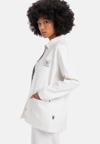 Veste blanche avec fermeture éclair, poches latérales et motif brodé sur la poitrine gauche. Tissu doux et texturé avec une coupe décontractée.