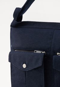 Granatowa torba na ramię z materiału, z kieszenią na klapce zabezpieczoną metalowym zatrzaskiem oraz kieszenią na zamek błyskawiczny powyżej.