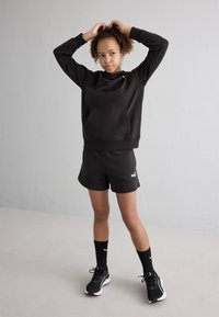 Sudadera con capucha negra y bolsillo frontal, combinada con pantalones cortos negros con un logo y zapatillas negras con detalles blancos.