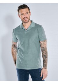 Green polo shirt with a textured fabric, short sleeves, and a collar. Worn with dark blue jeans. Tattoos visible on the left arm.