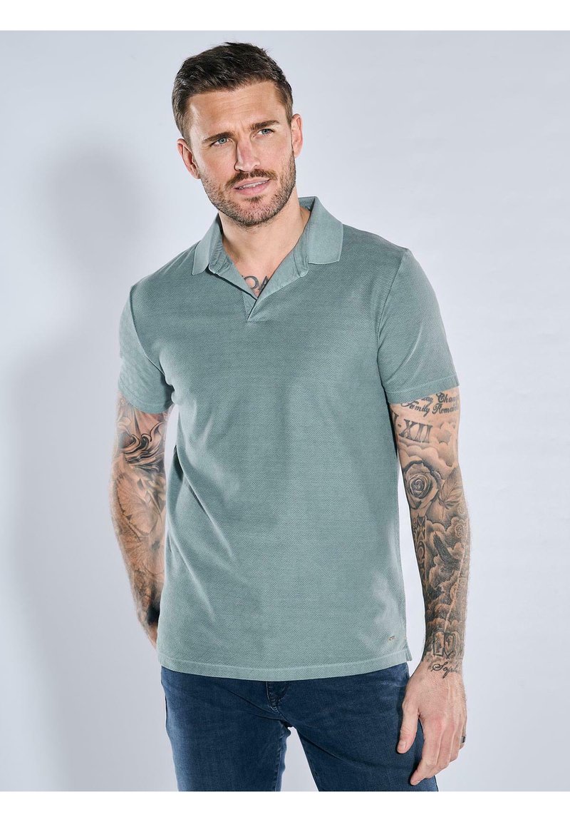 Green polo shirt with a textured fabric, short sleeves, and a collar. Worn with dark blue jeans. Tattoos visible on the left arm.