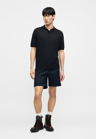 Polo noir à manches courtes associé à un short bleu marine, avec une coupe classique. Bottines marron à lacets avec une finition texturée et chaussettes blanches.