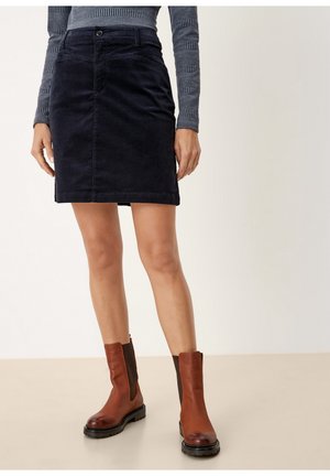 Vrouw draagt een zwarte rok tot aan de knie, een grijs patroon lange mouwen top en bruine leren enkellaarsjes, staand op een beige vloer.