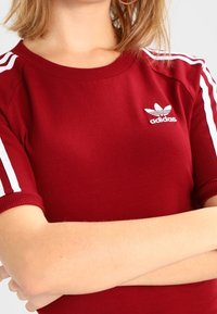 Czerwony sportowy top z prążkowanym okrągłym dekoltem, białymi akcentami w trzy paski na rękawach oraz małym białym logo Adidas na klatce piersiowej.
