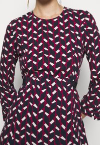 Sukienka z długim rękawem z geometrycznym wzorem w kolorach granatowym, białym i magentowym. Projekt zawiera jednokolorowe akcenty i dopasowaną talię.