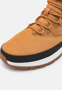 Brun mocka ovandel med en texturerad finish, med orangea accenter, svart gummisula och vit mellansula. Snörningssystem integrerat i designen.