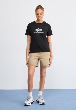 Black cotton t-shirt featuring a white Alpha Industries logo, paired with beige denim shorts and gray sneakers with black soles.