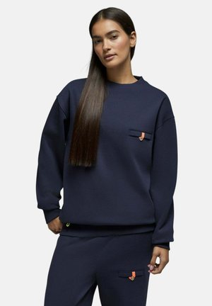 Femme aux longs cheveux raides portant un sweat-shirt bleu marine et un pantalon assorti avec des détails de poche orange.