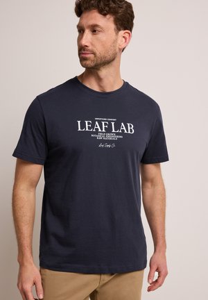 Homme aux cheveux bruns courts portant un T-shirt bleu marine avec l'inscription "LEAF LAB" et du texte botanique, associé à un pantalon beige, regardant vers la gauche, fond neutre.