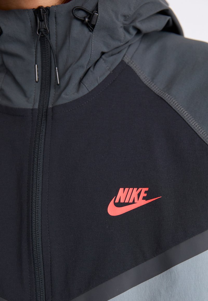 Kurtka Nike z zapięciem na zamek z przodu, kapturem i wzorem w kolorze czarnym i szarym, z wyraźnym czerwonym logo na piersi.