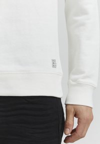 Felpa bianca in cotone con polsini e orlo a costine, dotata di un piccolo tag di marca sul lato inferiore, abbinata a pantaloni scuri e testurizzati.