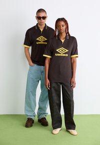 Čierne polo tričká s žltými akcentmi a logom "UMBRO", v kombinácii s ležérnymi džínsami a vzorovanými nohavicami, umiestnené na svetlom pozadí.