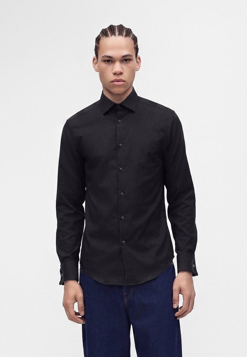 Jeune homme aux cheveux tressés portant une chemise noire ajustée boutonnée et un jean bleu foncé, debout devant un fond blanc uni.
