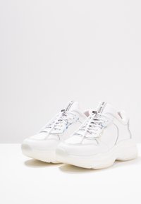 Zapatillas blancas voluminosas con un upper de cuero suave, acentos reflectantes y una suela gruesa texturizada. Los cordones y la marca en la lengüeta son visibles.