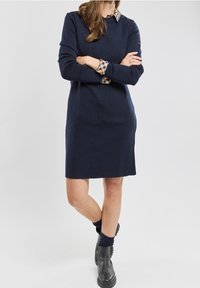Femme portant une robe bleu marine à manches longues sur une chemise à carreaux, avec des bottines noires et des chaussettes sombres, debout, les bras croisés.