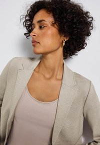 Blazer beige claro con solapas de muesca, tejido texturizado, llevado sobre una camiseta sin mangas gris claro, con una vista lateral en primer plano del portador.