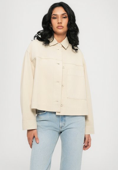 Beige cropped jacket with a pointed collar, front button closure, and a chest pocket. Smooth fabric with subtle stitching details.