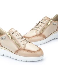 Zapatillas de cuero beige con cierre de cordones y cremallera, adornadas con acentos de tachuelas, una suela de goma blanca y una textura suave.