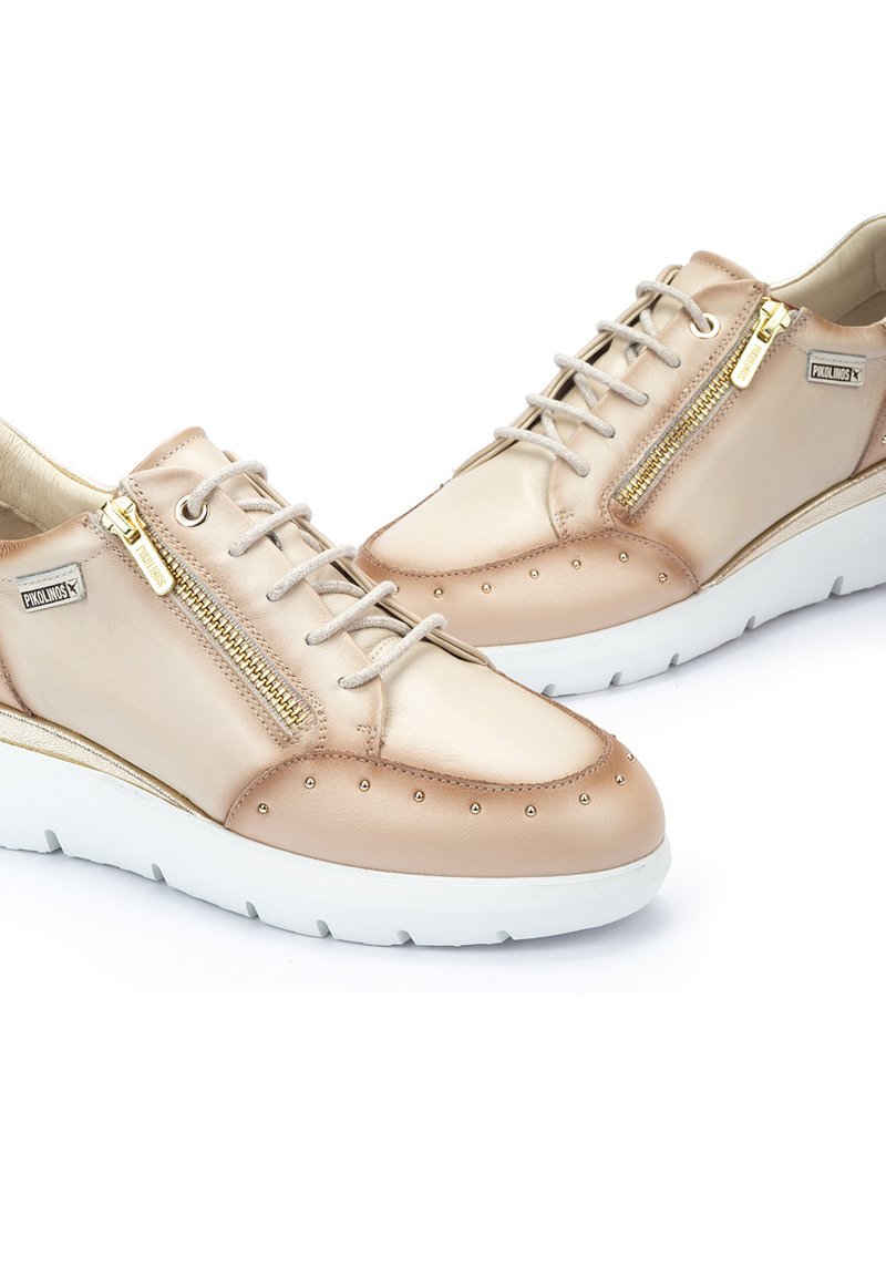 Zapatillas de cuero beige con cierre de cordones y cremallera, adornadas con acentos de tachuelas, una suela de goma blanca y una textura suave.