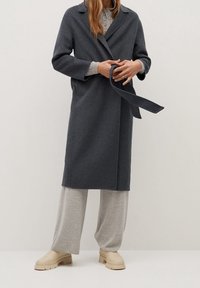 Personne portant un manteau gris foncé ceinturé sur un pull gris clair boutonné et un pantalon large, associé à des bottes beiges à semelle épaisse.