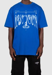 Modrá bavlněná trička s nápisem "ZTRACENÁ JUNGT" v bílém vlněném písmu. Volný střih s krátkými rukávy. Kombinováno s černými cargo kalhotami.