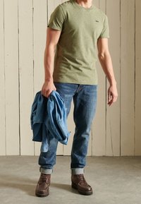 Grünes meliertes T-Shirt, blaue Denim-Jeans mit hochgekrempelten Säumen, braune Lederstiefel. Hält eine blaue Denim-Jacke. Neutraler Hintergrund.