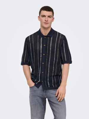 Giovane uomo che indossa una camicia blu navy a maniche corte con bottoni e righe verticali e jeans grigio chiaro, in piedi con una mano in tasca.