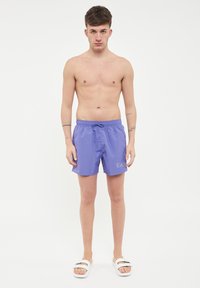 Pantaloni da bagno viola con vita elastica, coulisse frontale e logo stampato in basso a sinistra. Indossati con sandali bianchi a scorrimento.