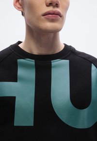 Μαύρο φούτερ με μεγάλο τιρκουάζ ανάγλυφο λογότυπο "HU" στο μπροστινό μέρος. Διαθέτει στρογγυλή λαιμόκοψη και υφασμάτινη υφή, με ριμπ μανσέτες.