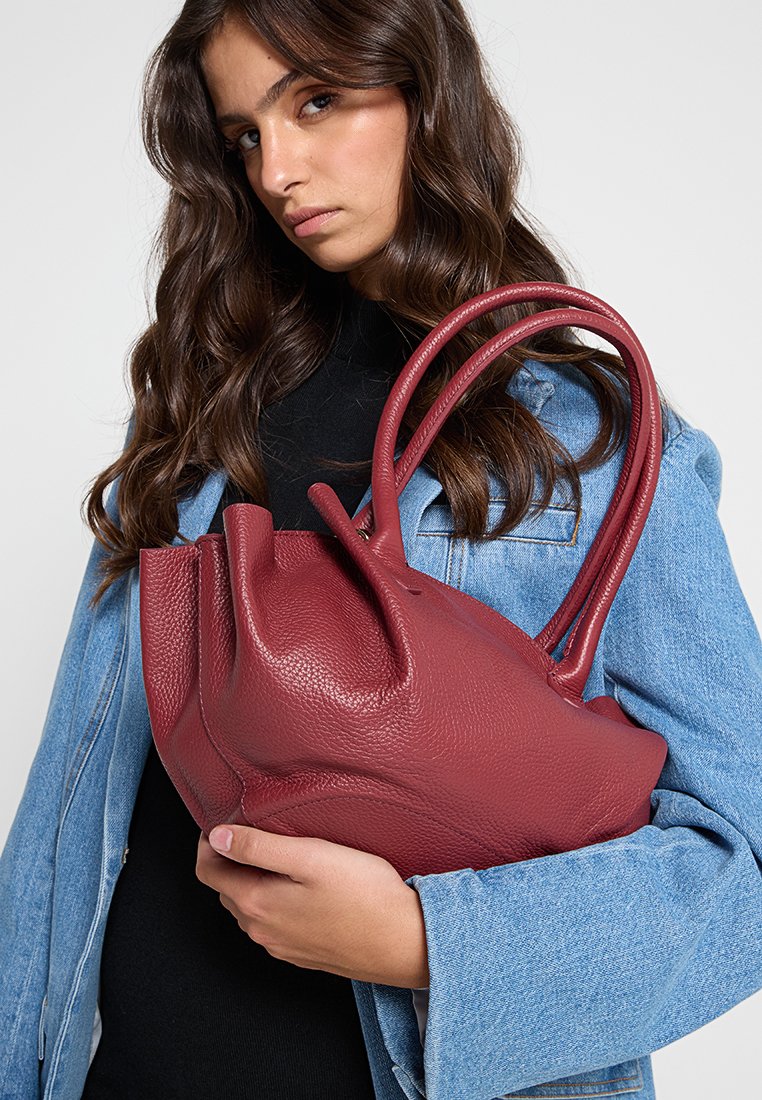 Borsa a tracolla rossa in pelle con superficie texturizzata, dotata di manici arrotondati e una forma scultorea unica. Indossata con un giubbotto di jeans.