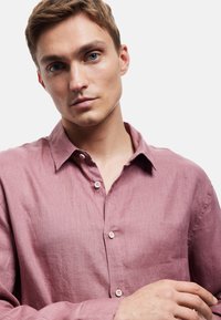 Camisa de lino rosa claro con cuello de botón, que presenta una textura visible y cuatro botones a lo largo de la tapeta.