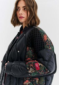 Czarna pikowana kurtka bomberka z kwiatowymi rękawami, siateczkowymi wstawkami i zapięciem na napy. Lewy rękaw ma czerwoną metkę.
