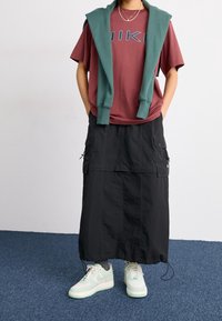 Person med en vinröd grafisk t-shirt, svart cargomaxikjol, mintgröna sneakers och en teal-färgad sweatshirt över axlarna.