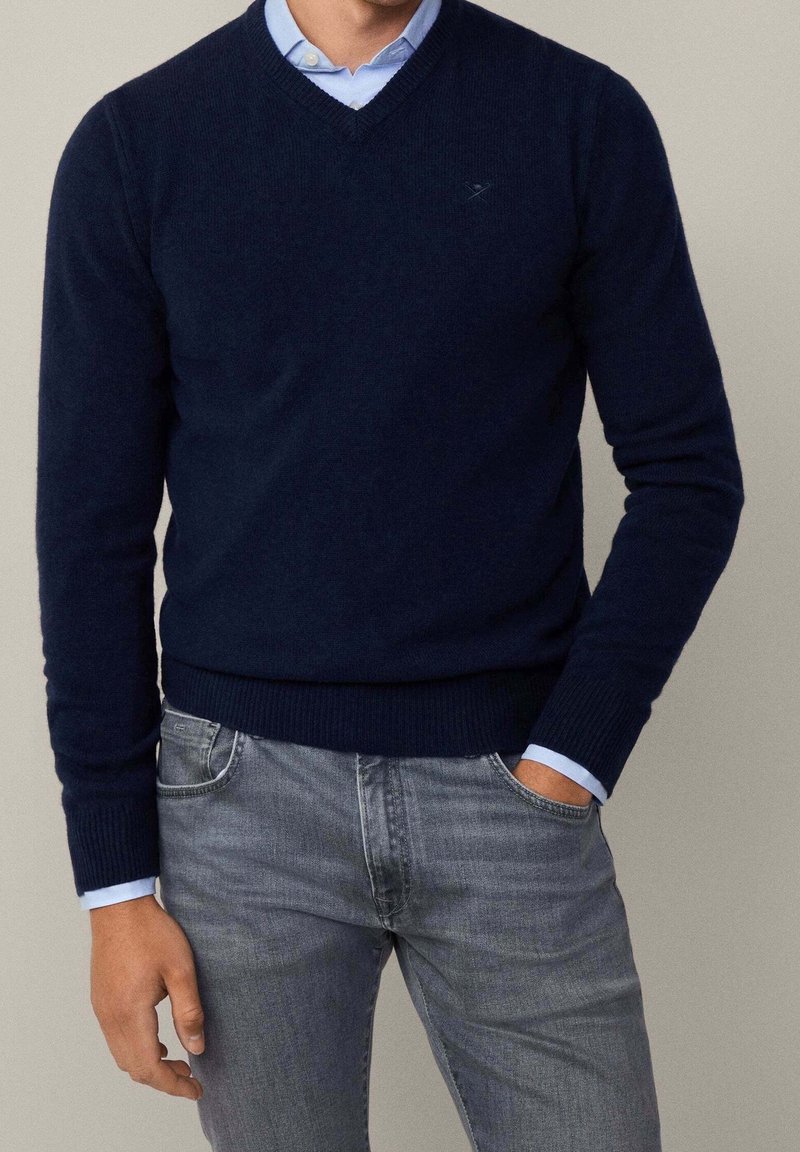 Homme portant un pull bleu marine à col en V par-dessus une chemise bleu clair avec un jean gris, debout avec une main dans la poche devant un fond uni.