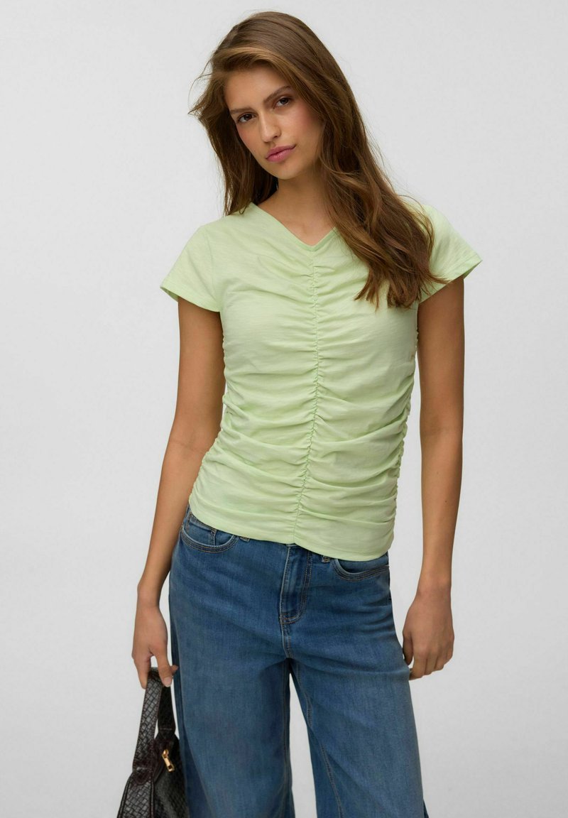 Jeune femme aux longs cheveux bruns portant un haut vert clair froncé à manches courtes et un jean bleu, tenant un sac à main noir texturé.