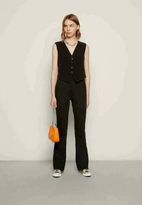 Frau in schwarzem Weste und Hose, die eine kleine orangefarbene Handtasche hält und vor einem schlichten hellen Hintergrund steht.