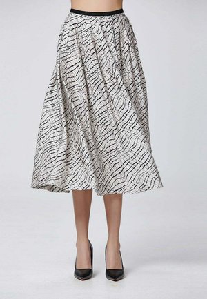 Femme portant une jupe blanche mi-longue avec des lignes abstraites noires et des chaussures à talons noirs, debout devant un fond uni.