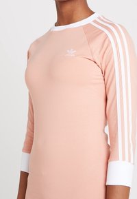 Kvinna som bär en ljusrosa Adidas-tröja med vita 3-randiga ärmar, vit krage och manschetter, med Adidas-logotypen på bröstet.