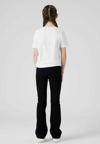 Weißes kurzärmliges T-Shirt, kombiniert mit schwarzen Schlagjeans und weißen Sneakers, von hinten gesehen, mit glattem braunem Haar in zwei Zöpfen.