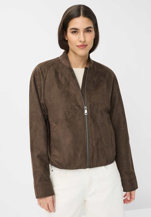 Woman with dark hair wears a brown suede zip-up jacket and white pants, standing against a plain light gray background.