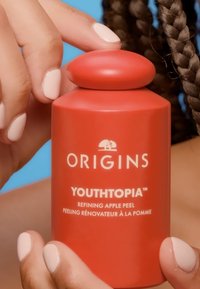 Bouteille cylindrique rouge avec un bouchon arrondi, étiquetée "ORIGINS YOUTHTOPIA Refining Apple Peel" en texte blanc. Surface lisse mate.
