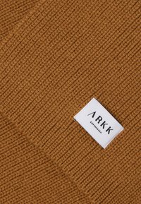 Bruin gebreide stof met een geribbelde textuur. Een wit label met "ARKK COPENHAGEN" in zwarte tekst is zichtbaar in de hoek.