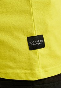 Ryškiai geltona medvilninė marškinė su juodu audinio etiketė, ant kurios siuvinėti žodžiai "ROCAWEAR" ir "ĮKURTA 1999".