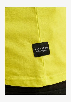 Fényes sárga pamut póló, melynek oldalvarrásába fekete szövetcímke van belevarrva, rajta a "ROCAWEAR" és az "ESTABLISHED 1999" feliratokkal.