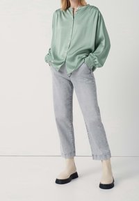 Blusa di raso verde con maniche a sbuffo, chiusura a bottoni; abbinata a jeans grigi chiari e stivaletti beige con suole nere.