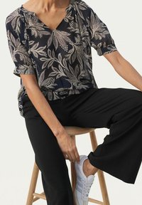 Blusa blu navy con motivi a foglia bianchi, maniche corte e scollatura a V, abbinata a pantaloni neri a gamba larga. Modella seduta su uno sgabello di legno.