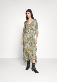 Grünes Maxikleid mit Blumenmuster, V-Ausschnitt, 3/4-Ärmeln und gerafftem Saum; kombiniert mit schwarzen Absatzstiefeln mit Schnallen.