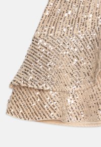 Tissu beige sequiné avec un ourlet à volants. Présente des rangées de sequins argentés brillants disposés en motif diagonal. Texture douce.